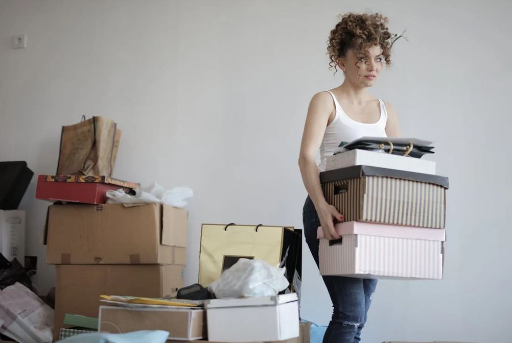 A woman holding and moving a couple of boxes wearing a white top and blue jeans